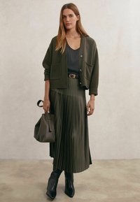 Maslinasto zeleni kardigan s džepovima, slojevit preko tamne majice, upareno s pliseranom maslinastom suknjom, crnim remenom i strukturiranom torbom. Crne gležnjače.