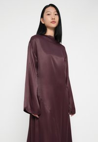 Burgundi sametkleit pikkade varrukatega ja kõrge kaelusega, pehme tekstuuri ja minimalistliku disainiga, mis langeb õrnalt külgedelt.