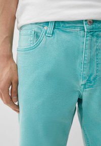 Turkusowe dżinsy o gładkiej strukturze, pięcioma kieszeniami i widocznymi szwami. Materiał ma lekki stretch dla zachowania komfortu.