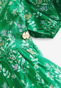Abito floreale verde con tessuto texturizzato, caratterizzato da piccoli fiori rosa e bianchi, e un bottone dorato con un design inciso.