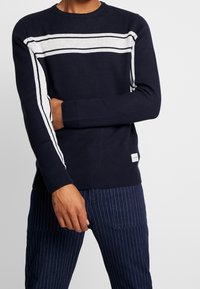 Navy gestrickter Pullover mit weißen Streifen, Rundhalsausschnitt und gerippten Bündchen. Strukturiertes Material und kleines Logoschildchen nahe dem Saum.