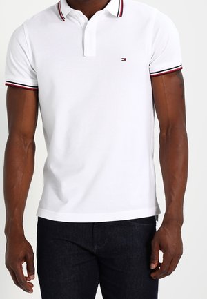 Mand iført hvid polo med rød og sort kant på krave og ærmer, kombineret med mørke jeans, stående foran en hvid baggrund.