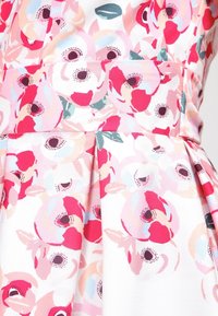 Robe fleurie avec un fond blanc orné de fleurs abstraites roses et rouges. Le tissu présente une texture lisse et des détails plissés à la taille.