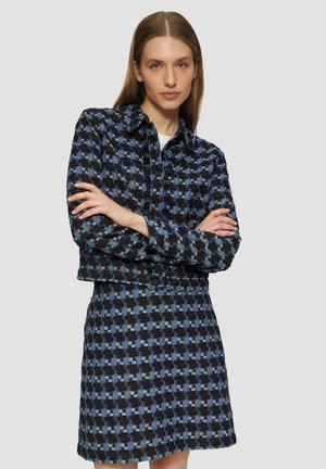 Blauw en zwart geblokt jasje en rokset, met een met kraag ontworpen, knoopsluiting aan de voorkant en een gestructureerde stof. Pose met gekruiste armen.