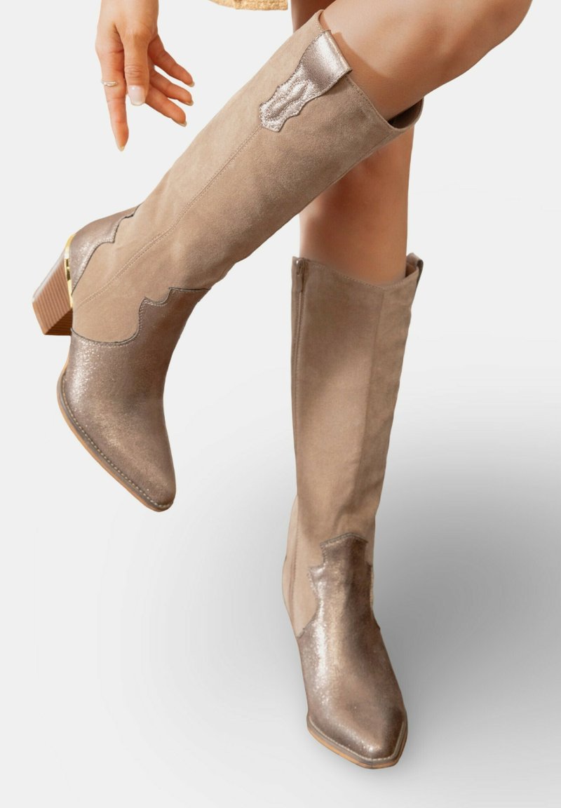 Bottes en suède beiges jusqu'aux genoux avec des accents métalliques, bout pointu, talon texturé et couture décorative sur le côté.