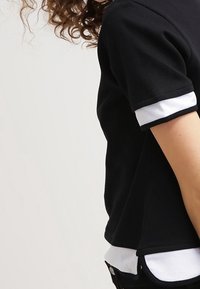 Osoba s kudrnatými vlasy oblečená v černém tričku s krátkým rukávem a bílým lemem na rukávech a spodním lemu, stojící na neutrálním světlém pozadí.