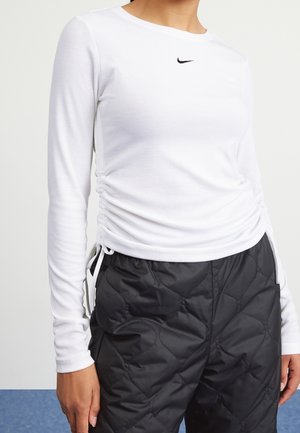 Frau trägt ein weißes, langärmeliges, gerafftes Nike-Oberteil und schwarze, gesteppte Hosen, steht mit einer Hand sichtbar an der Seite.