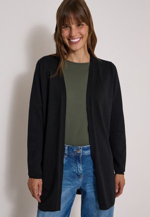 Femme souriante portant un cardigan noir ouvert, un haut vert et un jean bleu, debout devant un fond clair uni.