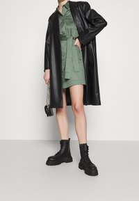 Persona che indossa un vestito corto verde con cintura, un lungo cappotto di pelle nera, stivali neri robusti con lacci e tiene una piccola borsa nera con una tracolla a catena.