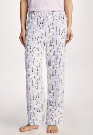 Woman wearing lavender pajama pants with white daisy flower print and a white drawstring, standing barefoot on a light background.