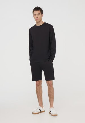 Set composto da una maglietta nera a maniche lunghe e pantaloni corti. La maglietta ha un colletto rotondo, mentre i pantaloni corti sono lunghi fino al ginocchio. Indossato con sneakers bianche e dettagli neri.