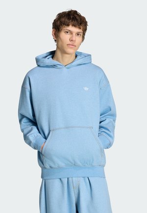 Jeune homme portant un sweat à capuche Adidas bleu clair et un pantalon assorti, les mains dans la poche avant, fond neutre.