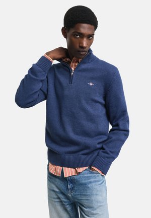 Suéter de punto azul con cuello con cremallera, puños acanalados y dobladillo, superpuesto a una camisa de rayas. Presenta un pequeño logo en el pecho.