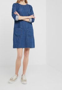 Tummansininen farkkuhenkseli, jossa on pyöreä kaula-aukko, kolmanneksen mittaiset hihat ja kaksi etutaskua. Yhdistetty vaaleansävyisiin tennareihin.