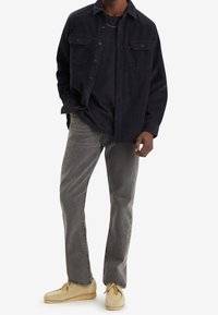 Mörk marinblå skjorta med knappar över en svart t-shirt, tillsammans med grå raka jeans och beigefärgade mockasinskor. Enkelt mönster, mjuk textur.