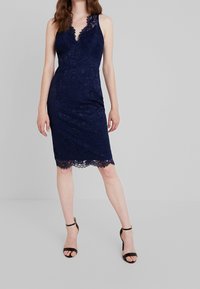 Vestido de encaje navy con escote en V profundo, cuerpo ajustado y dobladillo con forma ondulada. Presenta un patrón floral y tirantes ajustables. Combinado con sandalias negras.