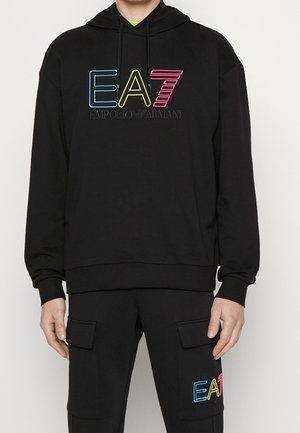 Personne portant un sweat à capuche noir et un pantalon avec des logos "EA7 Emporio Armani" colorés sur la poitrine et la jambe du pantalon.