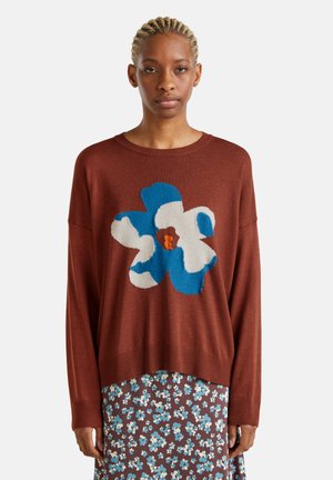 Brauner gestrickter Pullover mit einem großen blau-weißen Blumenmuster. Lockere Passform mit geripptem Saum. Kombiniert mit einem gemusterten braunen Rock.