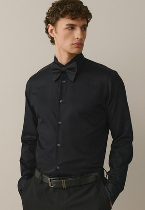 Young man wearing a fitted black dress shirt with a textured black bow tie, black trousers, and a black leather belt, looking to the side.