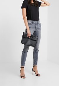 Bolso de mano negro de cuero con un asa superior, textura suave y forma rectangular, combinado con jeans ajustados grises y tacones negros de tiras.