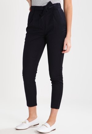 Femme portant un pantalon court noir à fines rayures taille haute avec une ceinture nouée et des mocassins blancs à enfiler, debout devant un fond uni.