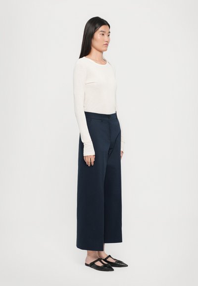 Beiger Langarmoberteil, marineblaue weit geschnittene Hose mit Knopfverschluss, schwarze spitze Mules mit geflochtenen Details, glatte Stoffstrukturen.