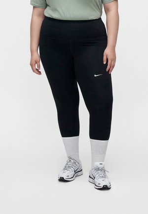Personne portant des leggings noirs Nike, des baskets blanches Nike et des chaussettes blanches, debout devant un fond clair uni.
