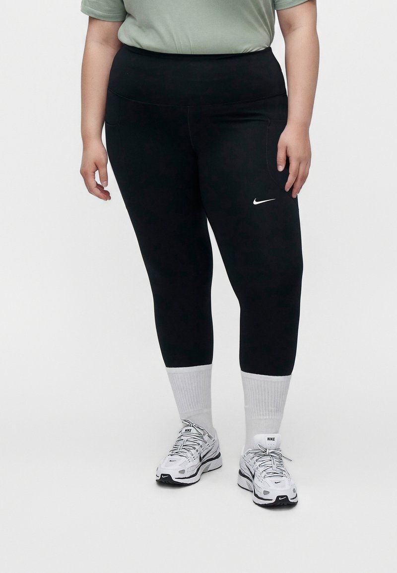 Personne portant des leggings noirs Nike, des baskets blanches Nike et des chaussettes blanches, debout devant un fond clair uni.