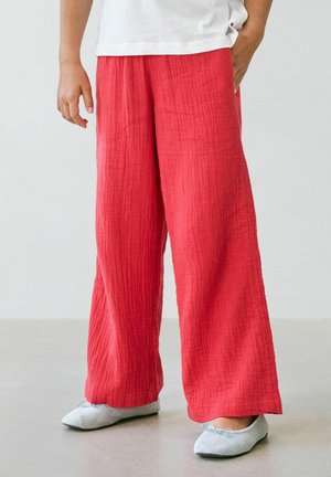 Person wearing loose, textured red pants, white top, and light gray ballet flats standing on a smooth floor.