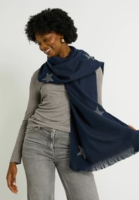 Écharpe marine en tissu doux, ornée d'étoiles argentées et avec des franges aux extrémités, drapée sur les épaules au-dessus d'un haut gris.