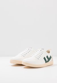 Białe trampki z zielonymi akcentami, zawiązywane, o gładkiej fakturze i beżowej gumowej podeszwie. Zawierają logo na języku i boku.