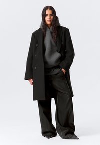 Γυναίκα με μακριά, σγουρά μαλλιά που φοράει μαύρο φαρδύ παλτό, γκρι φούτερ με κουκούλα, φαρδιά μαύρη παντελόνια και μαύρα μυτερά παπούτσια, όρθια μπροστά σε λευκό φόντο.