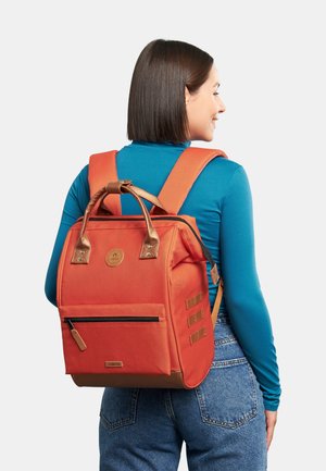 Sac à dos orange de forme rectangulaire, fabriqué en matériau durable. Dispose d'une poche zippée à l'avant, de poignées en bronze et de bretelles rembourrées.