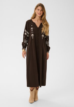 Femme portant une longue robe marron foncé avec broderies blanches de fleurs et d'oiseaux, détails de boutons, et bottines beiges sur un fond uni.