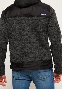 Hættetrøje i mørkegråt stof med en struktureret finish, med sorte nylon skuldre og et lille logo på ryggen.