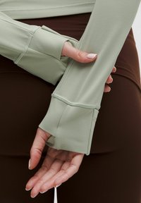 Light green long-sleeve top with thumbholes, featuring a smooth texture and flat seams. Paired with brown leggings, showcasing a simple design.