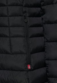 Zwarte pufferjacksectie met een geborduurde textuur, een ritszak en een klein logo-label in het rood aan de onderrand.