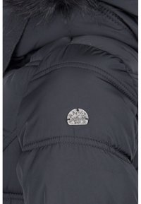 Sort dunjakkeærme med quiltet tekstur og en sølvmetallic label, der viser koordinater og et årstal, syet fast på stoffet.