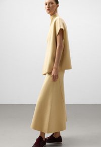 Donna in profilo che indossa una maglietta beige larga e una gonna lunga coordinata, con sneakers rosso scuro, su uno sfondo semplice.