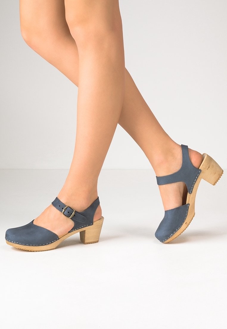 Personne portant des sabots en cuir bleu à talons avec des brides à la cheville et des semelles en bois, marchant sur une surface de couleur claire devant un fond uni.