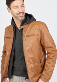 Brun læderjakke med en glat tekstur, der har to lynlåslommer på brystet, en opretstående krave og en grå hættetrøje lagdelt under.