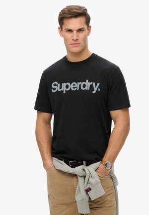 Black cotton t-shirt with "Superdry" logo in grey across the chest. Model wears khaki pants and a grey sweater tied at the waist.