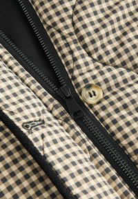 Veste matelassée avec un motif �à carreaux beige et noir. Elle dispose d'une fermeture éclair noire et d'un bouton crème, mettant en valeur des matériaux et textures contrastés.