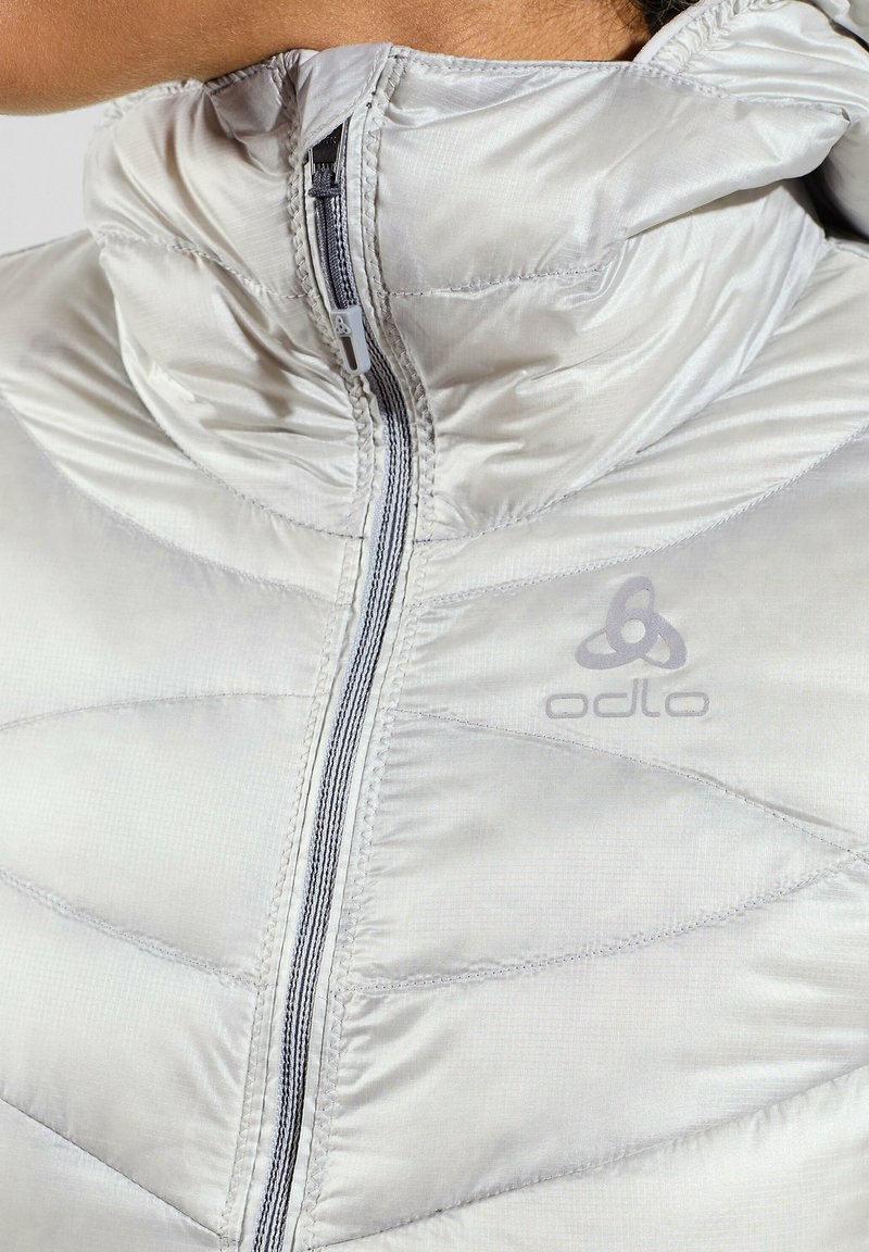 Hellgraue isolierte Jacke mit hohem Kragen, Reißverschluss, Chevron-Steppmuster und subtilen Branding-Details auf der Brust.