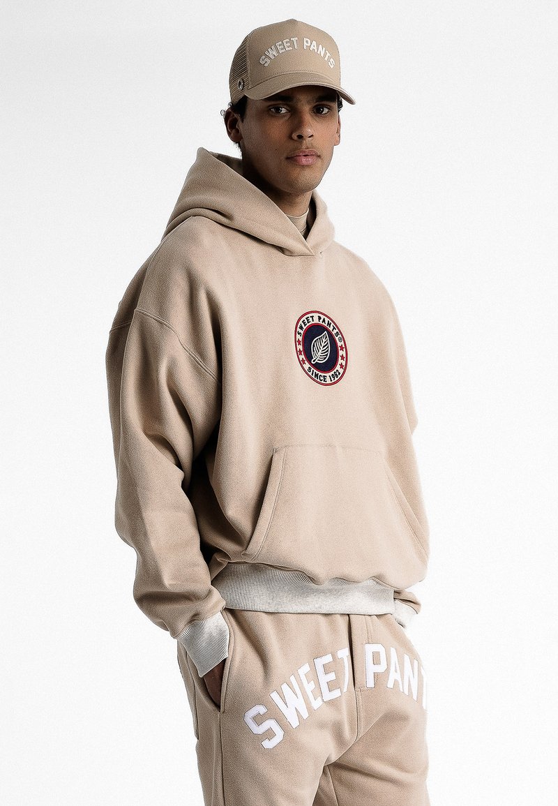 Sweat-shirt à capuche beige avec une poche avant et un logo brodé, associé à un pantalon de jogging assorti portant "SWEET PANTS" en lettres blanches.