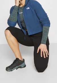 Blue zip-up hoodie with grey accents, black knee-length leggings, and black sports shoes with teal detailing. Textured fabric, athletic fit.