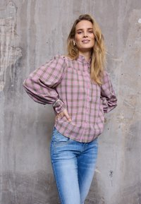 Femme aux longs cheveux blonds portant un chemisier à carreaux rose et un jean bleu clair, debout les mains sur les hanches devant un mur gris texturé.