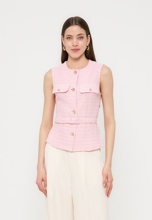 Mujer con chaleco sin mangas de color rosa texturizado con botones dorados y cinturón, combinado con pantalones anchos crema, sobre un fondo blanco.