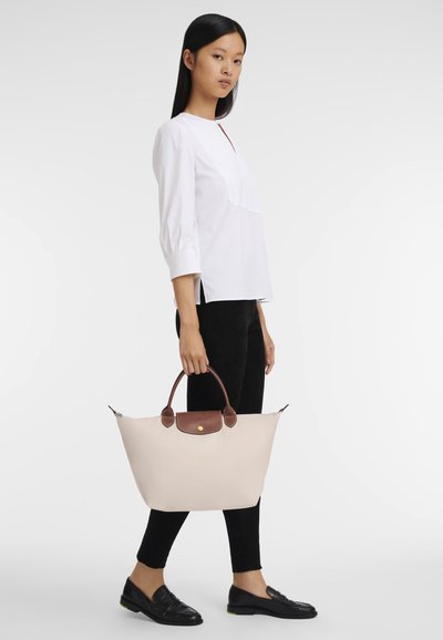 Beige tote bag with brown leather handle and accents. Smooth fabric, wide shape, and a zipper closure. Worn with a white top and black pants.