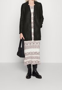 Svart ullkappa med rak skärning, ihop med en mönstrad krämfärgad och beige klänning samt svarta snörade kängor. Håller en svart väska.